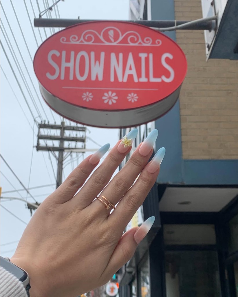 Ombre blue almond nails with a floral accent, photographed in front of the Show Nails salon sign